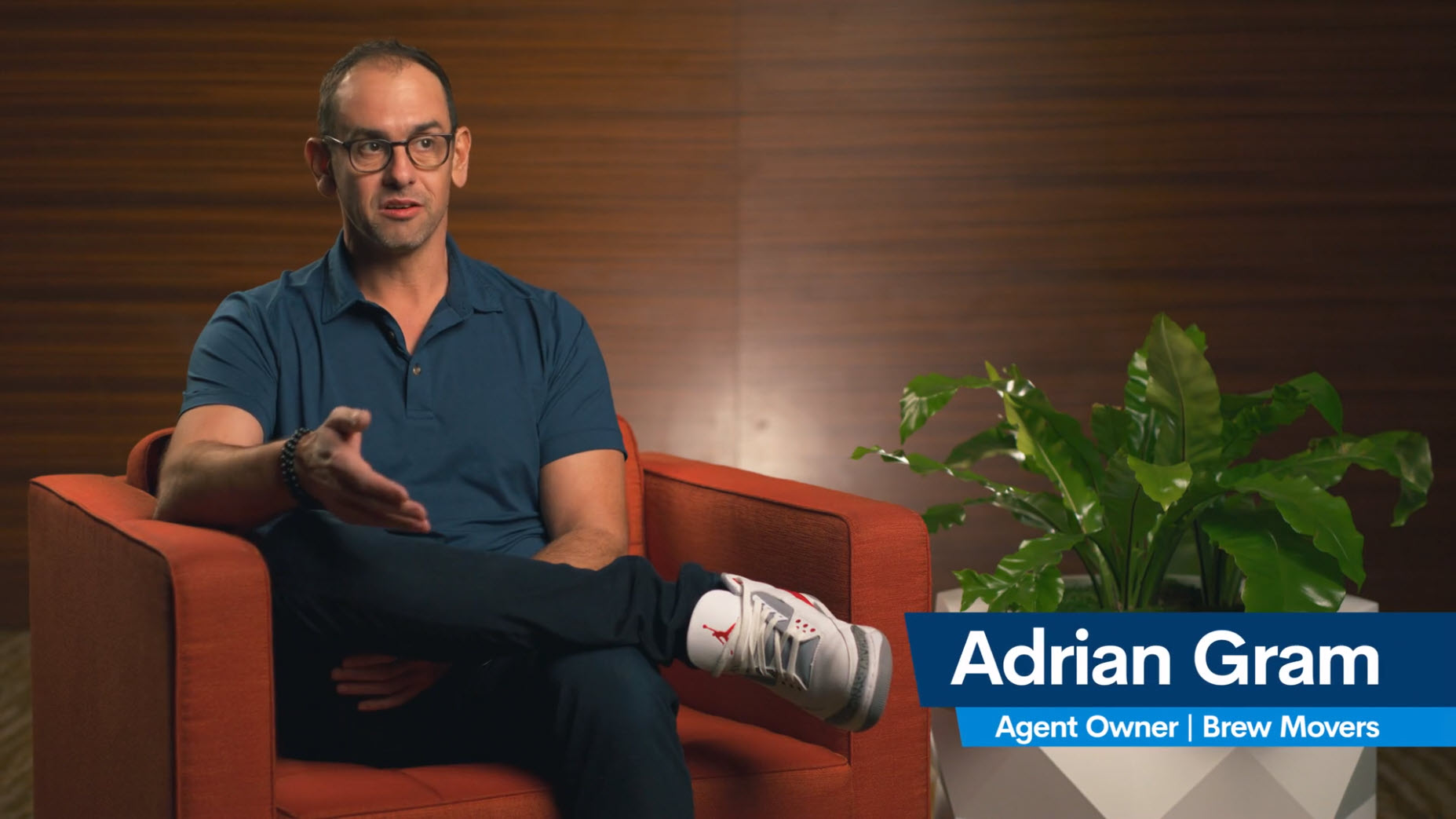 A person in a blue shirt and glasses sits in a red chair, gesturing with one hand. A green plant stands beside the chair. A text overlay reads: Adrian Gram, Freight Agent Owner | Brew Movers.
