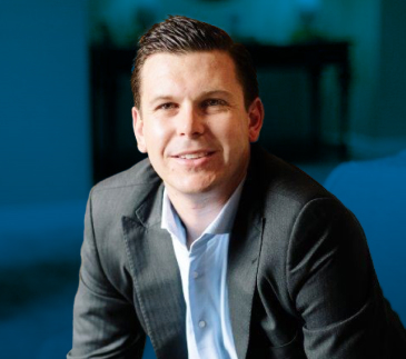 alex_stark A person in a dark suit and white shirt, exuding the confidence of a logistics agent, is smiling and looking at the camera. The background features a softly focused interior space with soothing shades of blue.