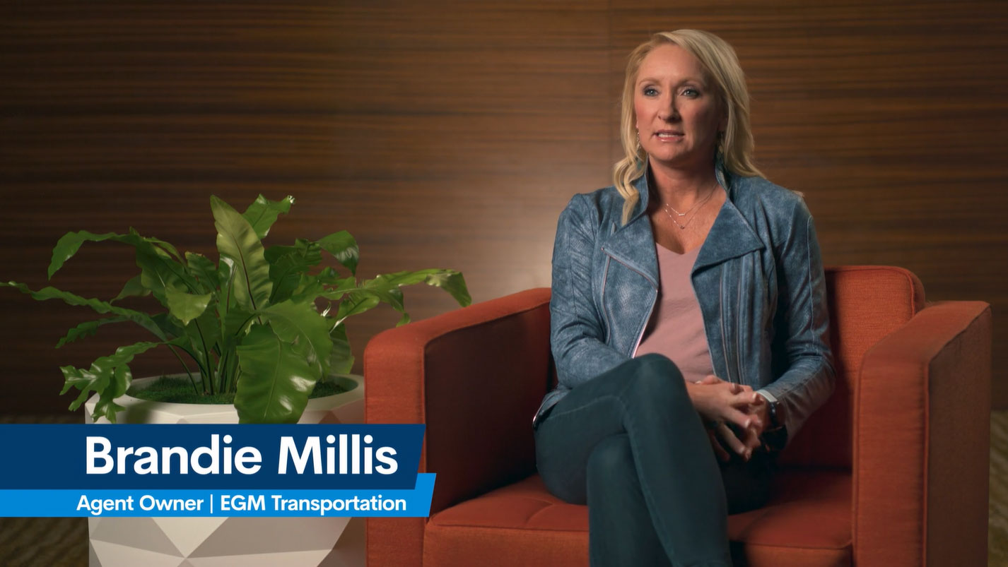 A woman sits in an orange chair beside a green potted plant, clad in a blue jacket and pink top. A text overlay reads, Brandie Millis, Agent Owner, EGM Transportation, hinting at freight agent opportunities. The backdrop is a wooden wall.