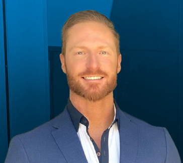 brian_coleman Man with short red hair and a beard, wearing a blue suit jacket over a dark shirt with a white collar, smiles confidently against a blue background, embodying the professionalism of a seasoned logistics agent.