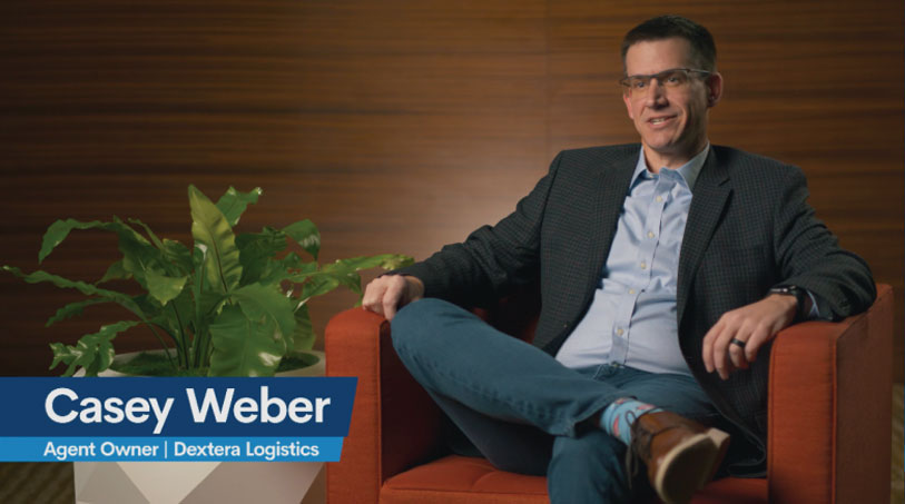 A man sits in an orange chair, wearing a blazer, jeans, and glasses. A green plant is beside him. The text reads Casey Weber, Independent Freight Agent Owner at Dextera Logistics. The background is a wooden panel wall.