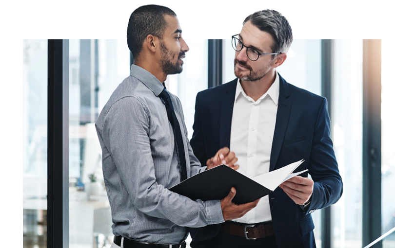 not-ready-cta Two men in business attire stand in an office, engaged in conversation. One, perhaps a seasoned freight agent, holds a folder open, pointing to a document, while the other listens intently. Both wear suits; one with glasses. Large windows frame the scene.