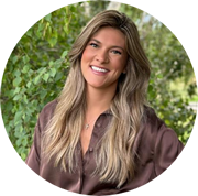 Smiling woman with long, wavy blonde hair wearing a brown button-up shirt stands outdoors in front of lush green foliage.