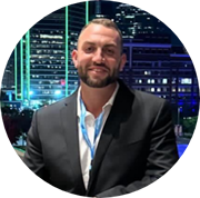 A man with short hair and a beard, wearing a dark suit and white shirt, stands smiling in front of a cityscape with brightly lit buildings at night.