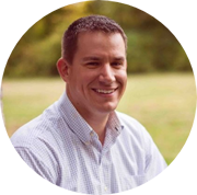 A man with short dark hair, wearing a light checkered shirt, is smiling while sitting outdoors with grass and trees in the background.