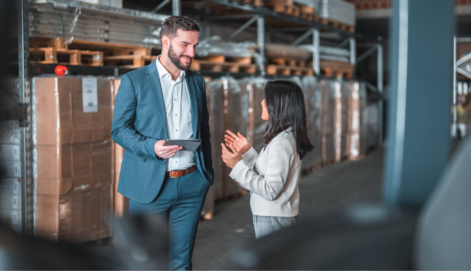 A man in a blue suit holding a tablet talks with a woman in business attire inside a warehouse with stacked pallets and cardboard boxes in the background.