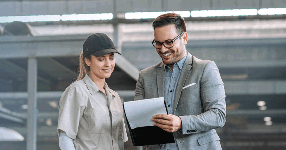 A man in a suit and a woman in a work uniform and cap are standing together indoors, smiling and looking at documents or a clipboard. The setting appears to be an industrial or warehouse environment.