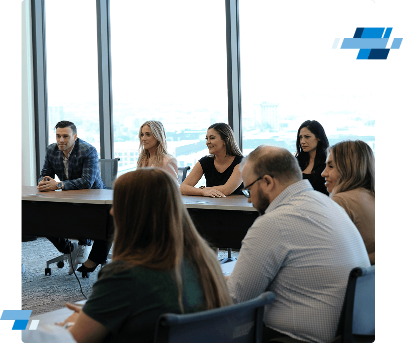 Freight Agent Culture4 A group of people sit at a conference table in a modern office with large windows, attentively listening to someone out of view. Some are smiling while others appear focused. Blue and white geometric graphics decorate the image corners.