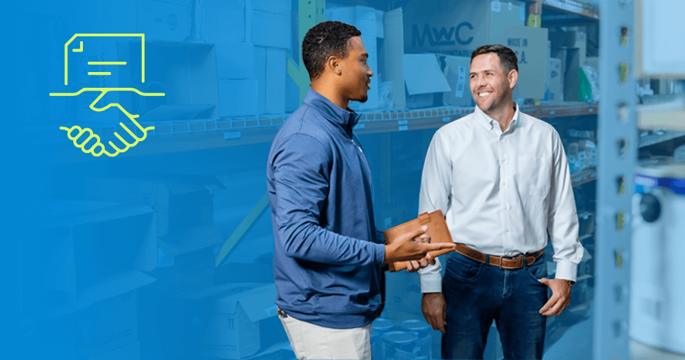 Two men standing and talking in a warehouse aisle. One holds a brown folder. They are smiling. On the left, a yellow graphic of a document above shaking hands is shown on a blue overlay. Boxes and shelves fill the background.
