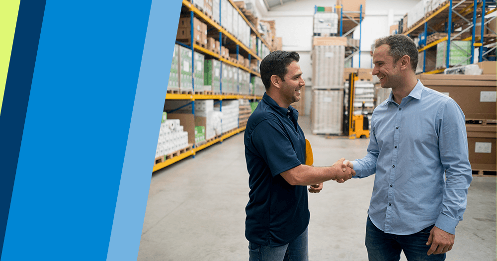 Two men smiling and shaking hands in a warehouse aisle, surrounded by shelves stocked with boxes and products—celebrating a successful partnership as one takes the next step to become a freight agent. One wears a navy polo, the other a light blue shirt.