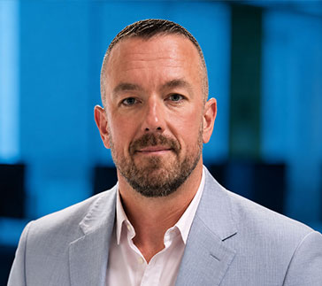 A man with short hair and a trimmed beard, embodying the professionalism of the GlobalTranz leadership team, wears a light gray suit jacket and white shirt as he stands in an office with a blue background.
